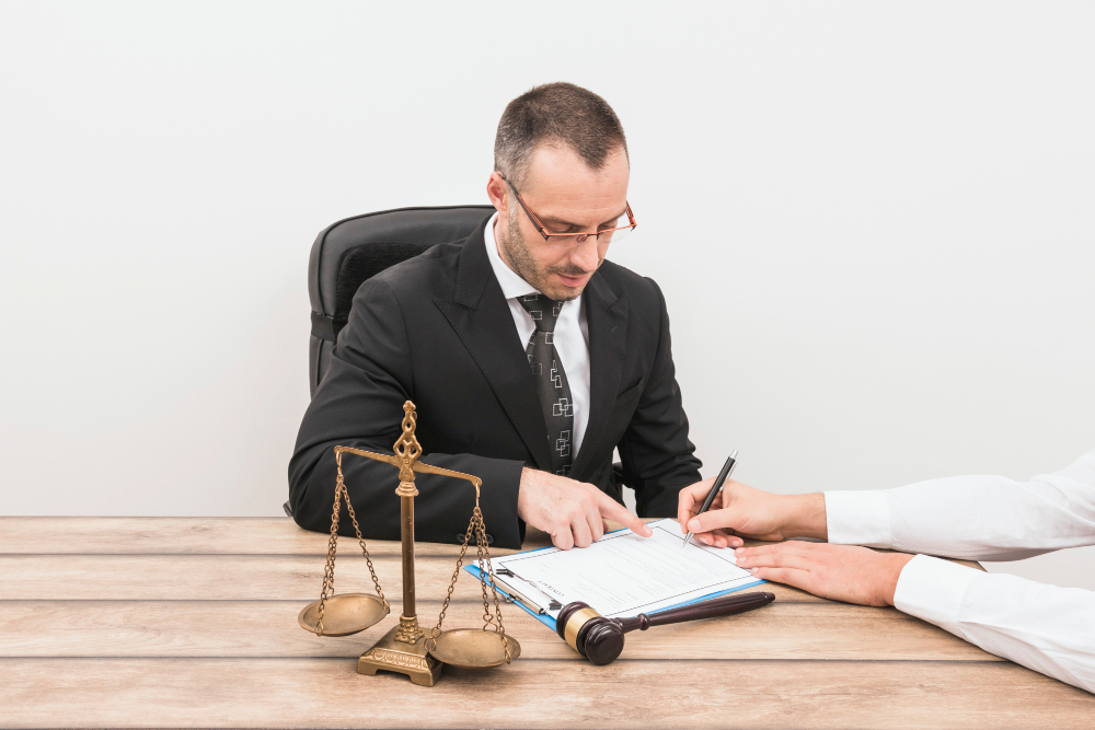 A lawyer reviewing and signing legal documents with a client, sitting at a desk with a gavel and justice scales in front.