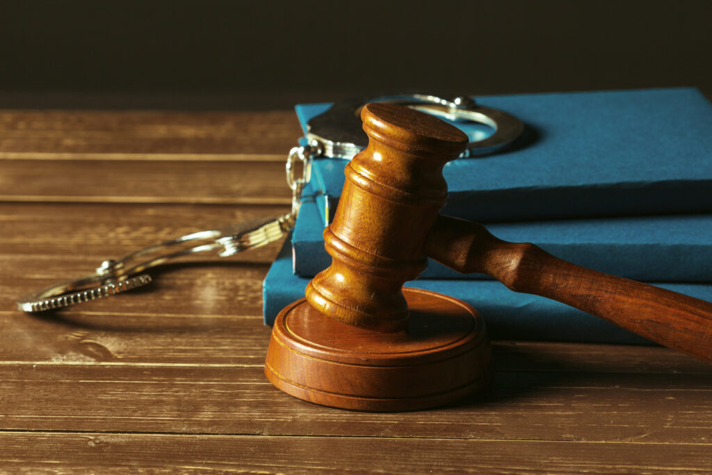 A wooden judge’s gavel resting on a sound block beside blue law books and a pair of metal handcuffs on a wooden table, symbolizing justice and criminal law.