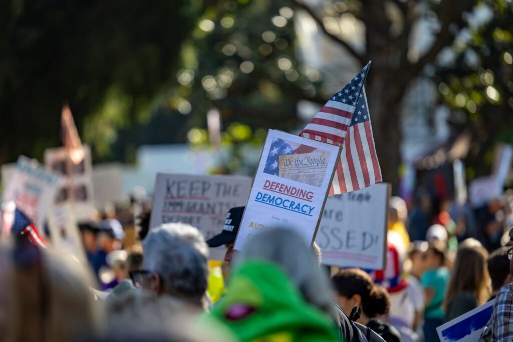 A diverse crowd holding an American flag and democracy signs, representing citizenship rights and protections under the 14th Amendment.